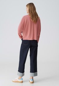 Hellt rosa, verkürzter Sweatshirt kombiniert mit dunklem Denim und aufgerollten Ärmeln. Die Schuhe haben einen weißen Körper mit pinken und blauen Streifen.