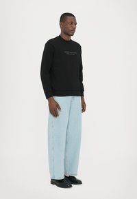 Juodas megztinis su "Armani Exchange" logotipu, derinamas su šviesiai mėlynomis plačiomis kelnėmis ir juodais batais, kuriuos dėvi stovintis modelis.