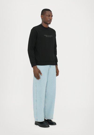 Sweatshirt noir avec le logo "Armani Exchange", associé à un jean large bleu clair et des chaussures noires, porté par un mannequin debout.