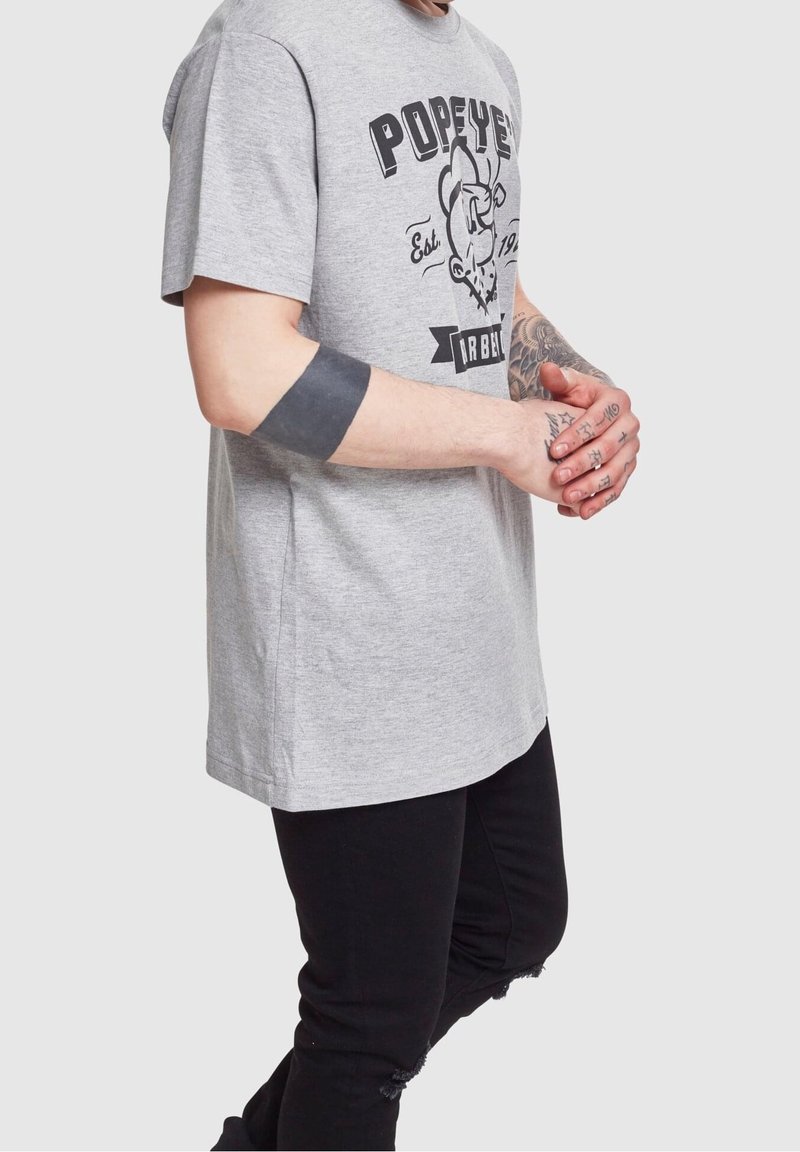 Camiseta de algodón gris con un estampado gráfico negro de Popeye. Corte relajado con mangas cortas. Combinada con jeans negros desgastados.