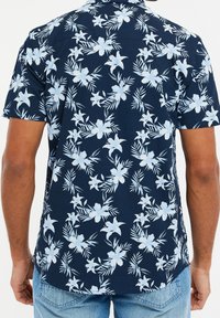 Chemise à manches courtes avec un fond bleu marine présentant de grands motifs de fleurs bleu clair et de feuillage vert. Le tissu a une texture lisse.