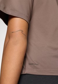 Kortermet, brun treningstopp laget av glatt stoff, med logodetalj i kanten og en avslappet passform. Hudtone synlig på armen.