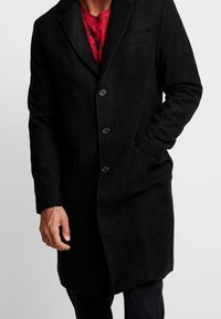 Cappotto in misto lana nero con collo a cono, tre bottoni, tasche laterali e una texture liscia. Caratterizzato da una silhouette sartoriale.