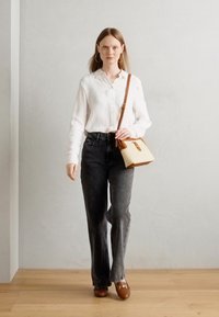 Modell i en hvit skjorte med knapper og mørke denim jeans med vidde, holder en kremfarget og brun skulderveske. Tregulv og grå veggbakgrunn.