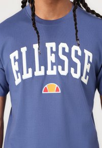 Mėlynas medvilnės marškinėlis su ryškiu baltu „ELLESSE“ tekstu ir spalvotu pusmėnulio logotipu apačioje. Apvalus kaklas ir trumpomis rankovėmis. Lygus paviršius.
