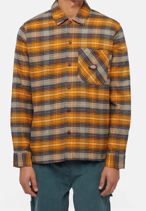 Buttoned long-sleeve orange, black, and cream plaid shirt with front chest pocket and logo patch worn with dark green pants.