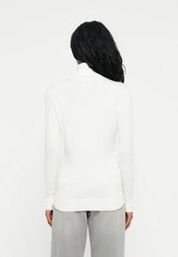 Persona con lunghi capelli neri che indossa un maglione bianco a collo alto e pantaloni grigio chiaro, in piedi di spalle rispetto alla fotocamera.