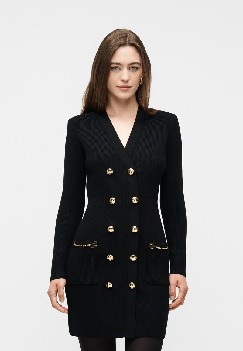 Γυναίκα με μακριά καστανά μαλλιά, που φοράει εφαρμοστό μαύρο φόρεμα με μακριά μανίκια, χρυσά κουμπιά και λεπτομέρειες αλυσίδας στις μπροστινές τσέπες, στέκεται μπροστά σε λευκό φόντο.