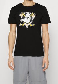 Man i svart t-shirt med en stiliserad vit hockeymask och korslagda gula hockeyklubbor, i kombination med ljusgrå shorts.