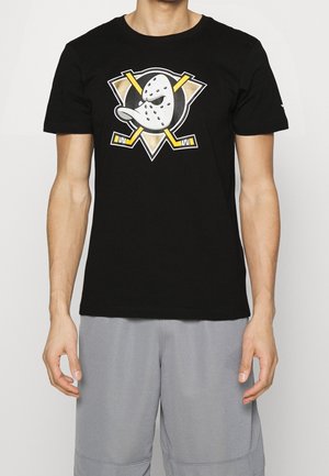 Homme portant un t-shirt noir avec un masque de hockey stylisé blanc et un graphique de crosses de hockey jaunes croisées, associé à un short gris clair.