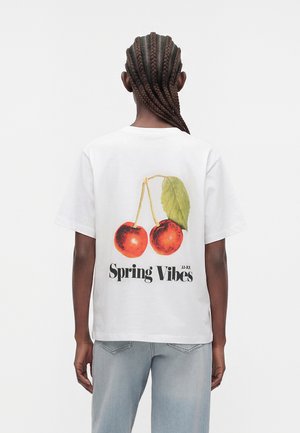 Osoba z warkoczami nosi białą koszulkę z czerwonymi wiśniami, zielonym liściem oraz napisem „Spring Vibes”, w połączeniu z jasnoniebieskimi dżinsami.