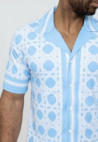 Chemise à manches courtes bleu clair présentant un motif géométrique blanc, un col pointu et une fermeture à boutons avec des rayures horizontales près des manches.
