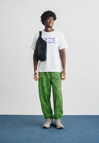 Μαύρη τσάντα χιαστί, λευκό μπλουζάκι με μωβ σχέδιο, πράσινο παντελόνι με μοτίβο και γκρι αθλητικά παπούτσια, στέκεται μπροστά σε φωτεινό φόντο.