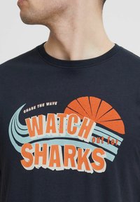 Navyblaues Baumwoll-T-Shirt mit einem grafischen Druck, der in orangefarbenem und hellblauem Text "WATCH out for SHARKS" und wellenförmigen Akzenten zeigt.