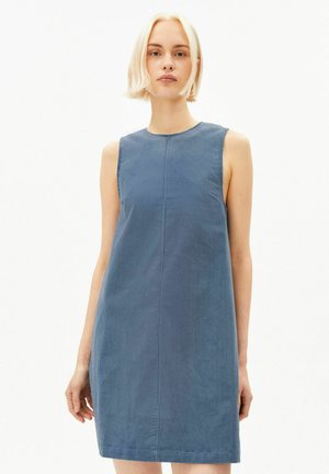 Junge Frau mit kurzen blonden Haaren, die ein ärmelloses blaues Cordkleid vor einem schlichten hellen Hintergrund trägt.