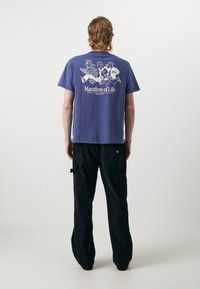 Camiseta de algodón morada de manga corta con una impresión blanca de tres figuras corriendo y el texto "Maratón de la Vida" en la parte posterior. Pantalones cargo negros.