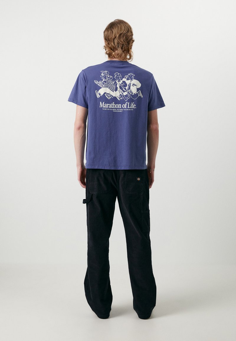Camiseta de algodón morada de manga corta con una impresión blanca de tres figuras corriendo y el texto "Maratón de la Vida" en la parte posterior. Pantalones cargo negros.