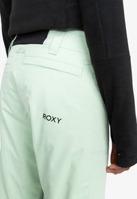 Myntegrønne bukser med fire lommer og sort "ROXY" logo på bagsiden. Teksturen ser glat ud, parret med en sort fleece-top.