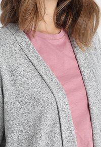 Cardigan grigio realizzato in un materiale a maglia testurizzato, con un'apertura frontale e una T-shirt rosa chiaro sotto, con una finitura liscia.