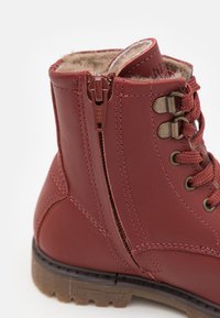 Bottine en cuir rouge avec doublure en fausse fourrure, cordon de serrage à l'avant et fermeture éclair sur le côté. Semelle en caoutchouc avec semelle texturée pour une meilleure adhérence.