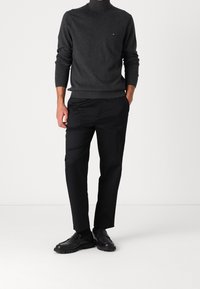 Mann in dunkelgrauem Rollkragenpullover, schwarzen Hosen und schwarzen Slippern, der mit Händen in den Taschen vor einem schlichten Hintergrund steht.