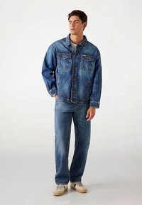 Geacă din denim albastru mediu, cu două buzunare pe piept, închidere cu nasturi și croială lejeră, asortată cu blugi albaștri și adidași.