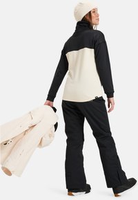 Pufferjacke in lichtcrème met een gestructureerde zwarte bovenkant, gecombineerd met zwarte sneeuwbroeken. Het model houdt een bijpassende crèmekleurige jas vast.