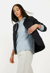 Gequilted zwarte jas met drukknoopsluiting, gelaagd over een lichtblauwe hoodie. Witte broek zichtbaar, benadrukt een casual en gelaagd outfitontwerp.