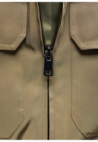 Khaki-Jacke mit Zentralreißverschluss, ausgestattet mit zwei Klappentaschen. Hergestellt aus strukturiertem Stoff mit einem glatten Reißverschluss und verstärkten Nähten.