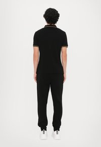 Tricou polo negru cu accente galbene, asortat cu pantaloni de trening negri. Modelul se află cu spatele, încălțat cu adidași albi cu dungi negre vizibile.