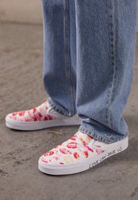 Baskets blanches à enfiler avec des motifs d'empreintes de lèvres en rouge et rose, avec le texte "LOVE CAN SAVE US" en noir sur le côté. À porter avec un jean bleu.