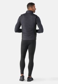 Schwarze isolierte Weste mit hohem Kragen, strukturiertem Vorderteil und glatten Seiten. Tragen über einem dunklen Langarm-Oberteil und eng anliegenden schwarzen Leggings.