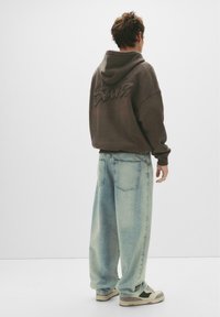 Hoodie marron avec un motif dans le dos, associé à un jean en denim bleu clair surdimensionné et des baskets beiges avec des accents verts.