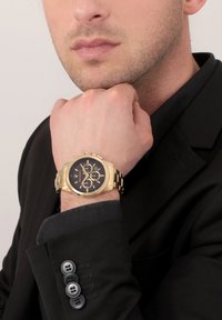 Orologio cronografo dorato con quadrante nero e tre sotto-quadranti, indossato su un polso, contro una giacca da completo nera con dettagli sui bottoni.