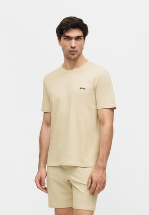 Mand iført beige kortærmet T-shirt med "BOSS"-logo og matchende shorts, stående foran en ensfarvet lys baggrund.