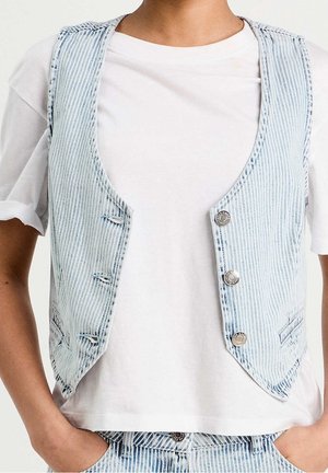 Personne portant un t-shirt blanc sous un gilet en denim rayé bleu clair avec des boutons en métal et un pantalon en denim rayé assorti.