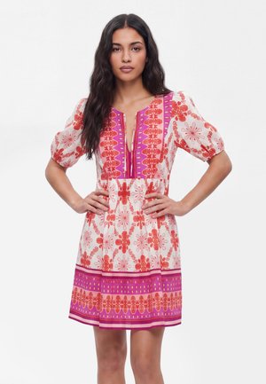 Mujer con cabello largo y oscuro que lleva un vestido corto blanco con estampados florales rojos y bordes decorativos rosas, parada con las manos en las caderas.