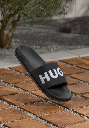 Einzelne schwarze Slide-Sandale mit weißem "HUG"-Schriftzug auf dem Riemen, positioniert über Holzblöcken mit kleinen Steinen dazwischen im Freien.