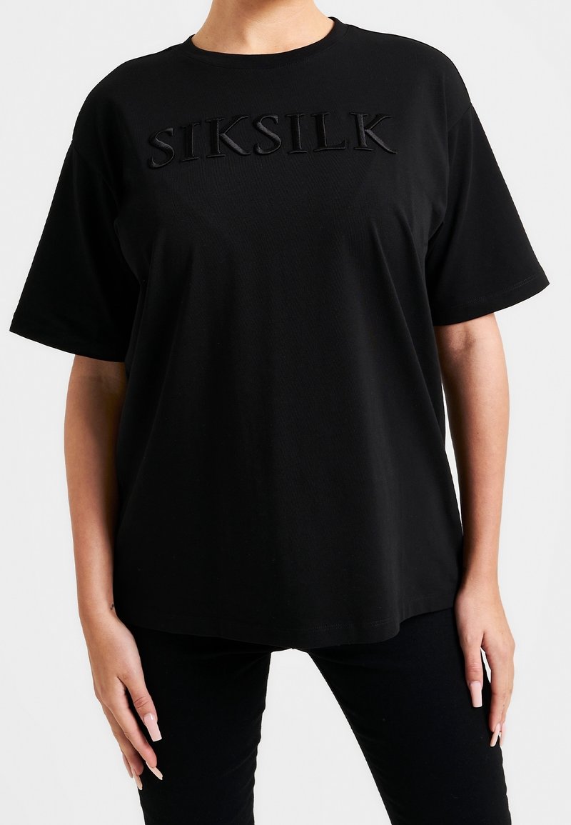 Osoba nosící černé oversized tričko s vyvýšeným černým nápisem "SIKSILK", v kombinaci s černými kalhotami, stojící na bílém pozadí.
