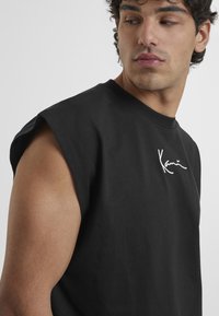 T-shirt noir oversized sans manches en coton, arborant un logo signature blanc sur la poitrine, avec un col rond et une coupe décontractée.