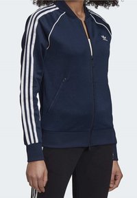 Osoba ve tmavě modré bundě na zip Adidas s bílými pruhy na rukávech a černých kalhotách, které mají bílé pruhy po stranách.