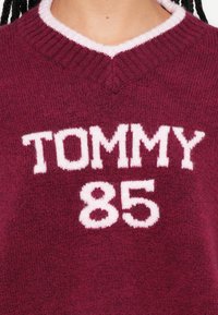 Burgundy värvi kampsun V-kaelusega, mille esiküljel on valgete kootud tähtedega "TOMMY 85". Pehme tekstuur ja soonikkoelised kaeluse detailid.