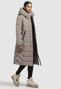 Žena kráča, má na sebe dlhý taupe prešívaný kabát s kapucňou, čierne legíny a béžové masívne šnurovacie čižmy na jednoduchom pozadí.