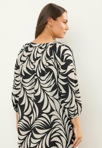 Sort og creme bluse med bladmønster, pufærmer, elastiske manchetter og en samlet halsudskæring. Bagsiden har en plissédetalje.