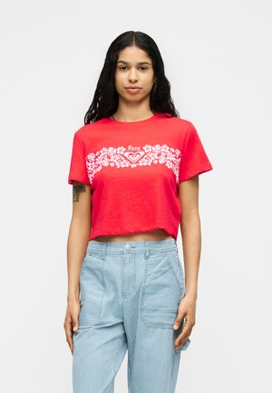 Femme aux longs cheveux foncés portant un t-shirt court rouge avec un motif floral blanc et un pantalon bleu rayé, debout devant un fond uni.