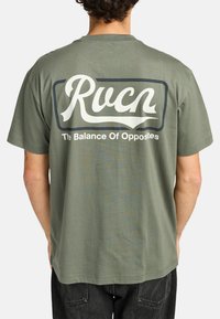Olivgrünes Baumwoll-T-Shirt mit kurzen Ärmeln. Weiße Schrift auf der Rückseite lautet "Rucn" mit "The Balance Of Opposites" darunter, umrahmt von einem Rechteck.
