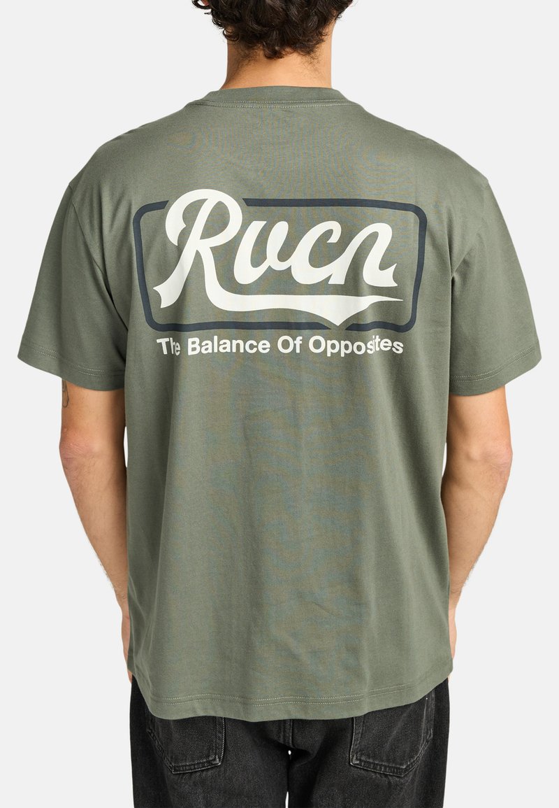 Olivgrünes Baumwoll-T-Shirt mit kurzen Ärmeln. Weiße Schrift auf der Rückseite lautet "Rucn" mit "The Balance Of Opposites" darunter, umrahmt von einem Rechteck.