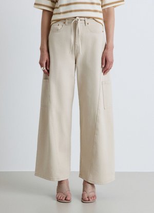 Persona che indossa pantaloni beige a gamba larga con coulisse in vita e sandali beige con punta aperta, in piedi su uno sfondo semplice.