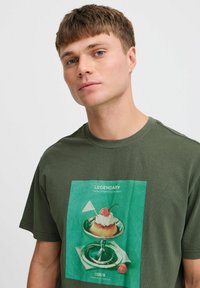 Camiseta de algodón verde oliva con un gráfico colorido de un postre en un plato plateado, con cerezas y una pequeña bandera.