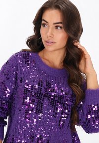 Lila Pullover, der mit Pailletten bedeckt ist, locker sitzt, einen gerippten Kragen hat und aus strukturiertem Strickstoff besteht. Verfügt über verschiedene Schattierungen von Lila.